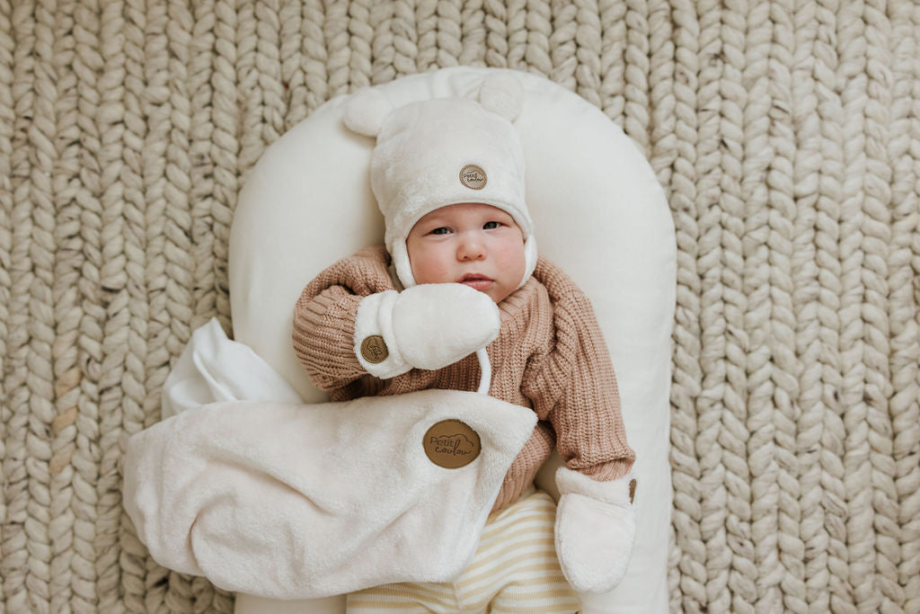 Bébé couché sur un coussin au sol portant ses accessoires d'hiver Petit Coulou dans la couleur crème