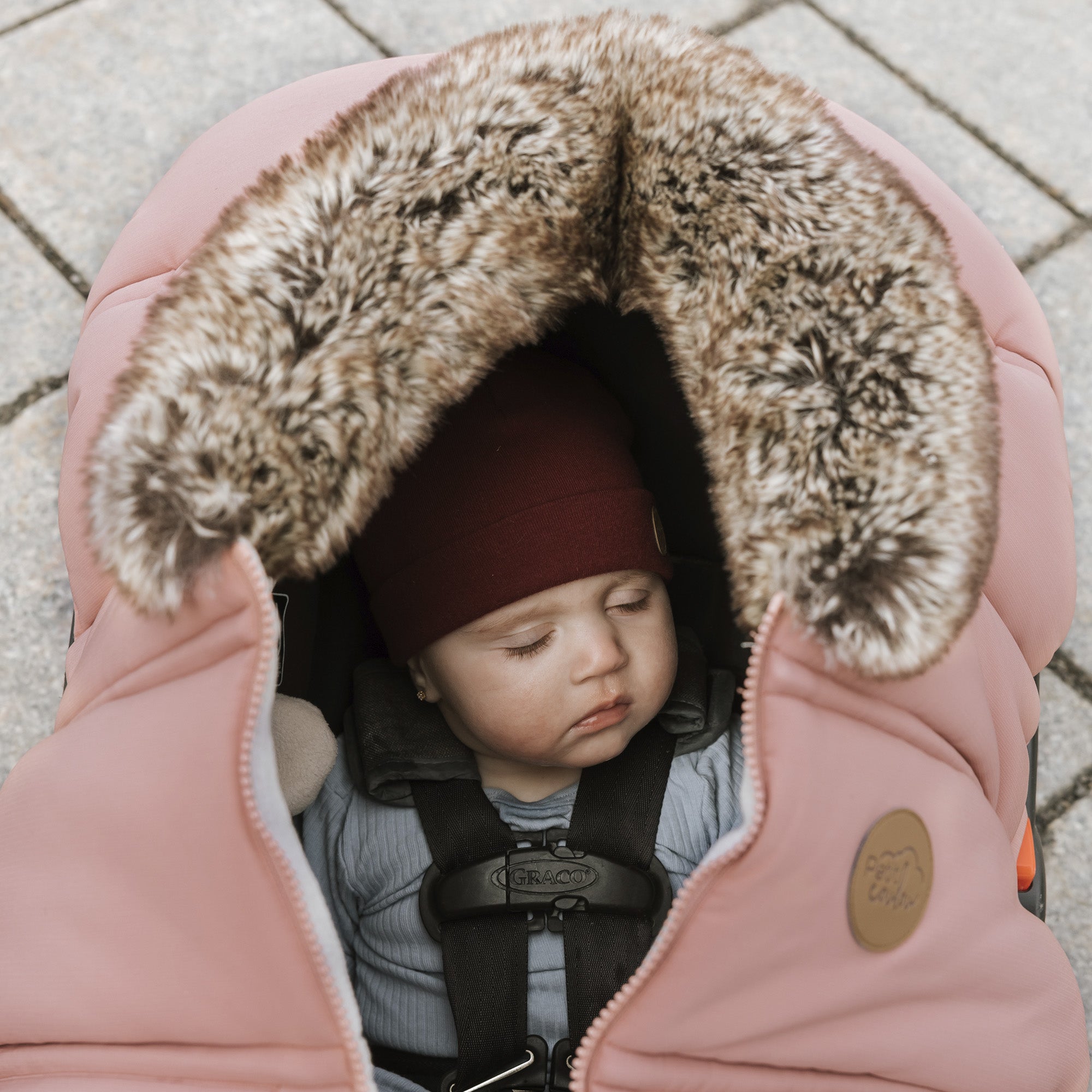 Bébé qui dort confortablement dans sa coquille recouvert d'une housse hiver classique Petit Coulou dans la couleur Blush avec sa tuque pompon rubis lors d'une marche automnale