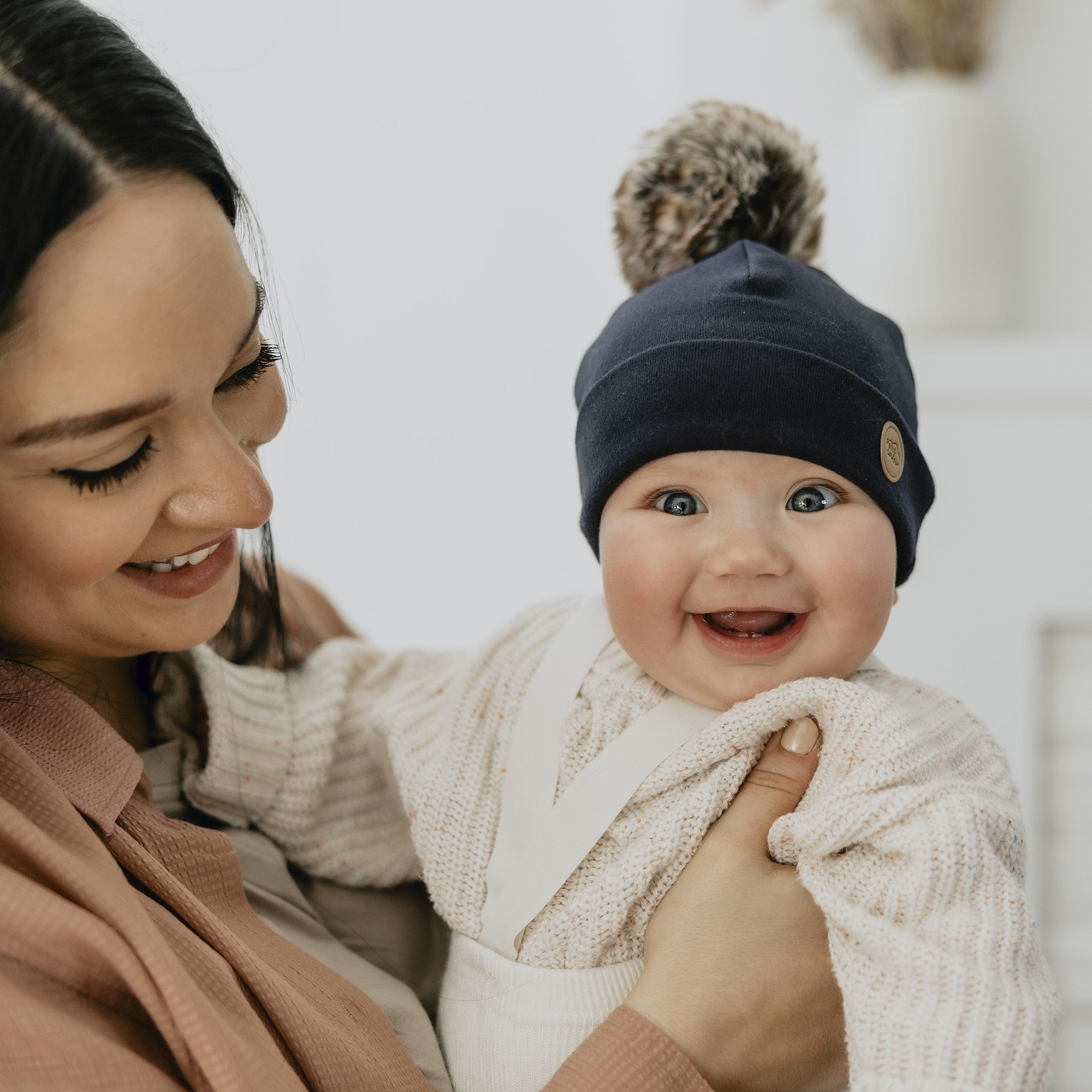 Bébé dans les bras de maman avec un gros sourire en regardant la caméra avec sa tuque pompon Petit Coulou dans la couleur agate