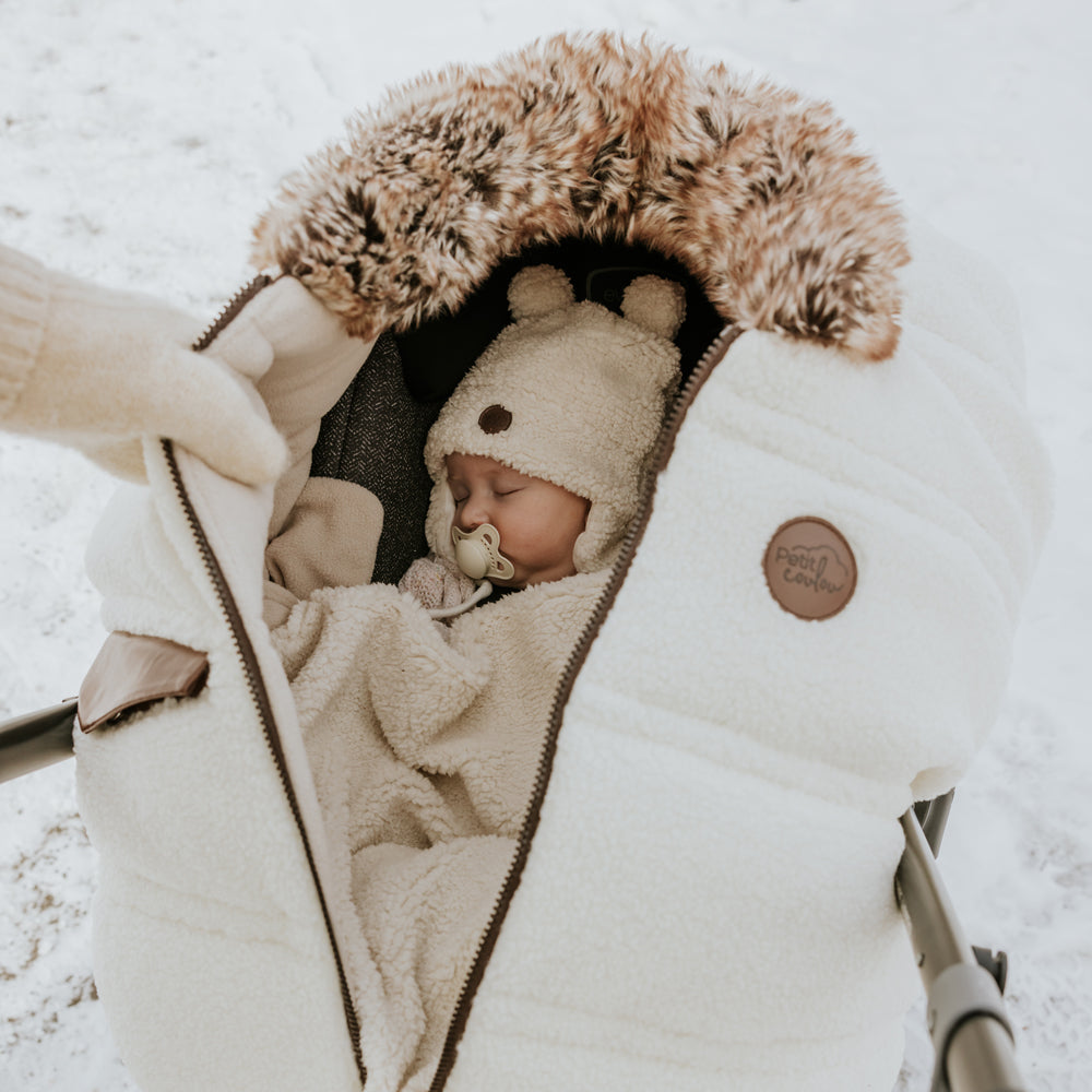 Little baby with a sherpa teddy bear hat and blanket in her car seat with a Jasper winter cover on it.