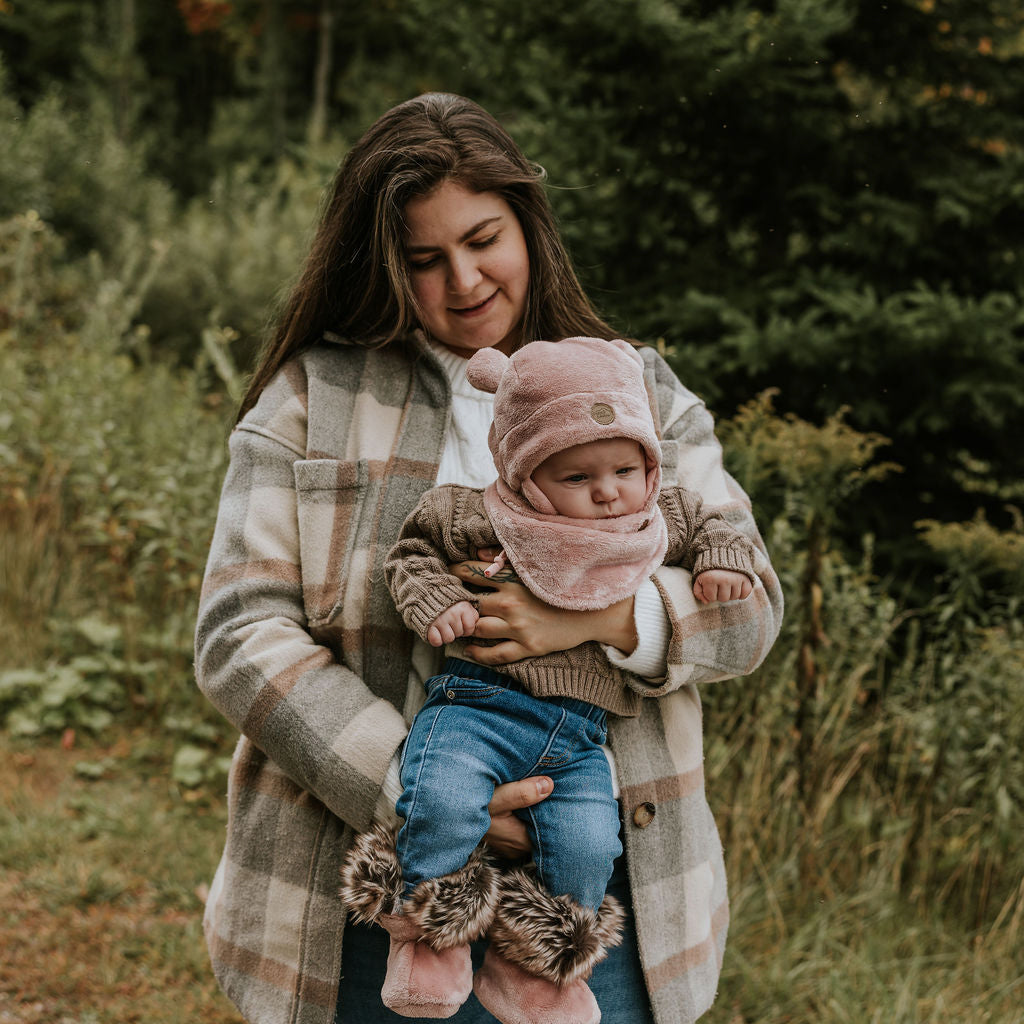 Mère qui tien son bébé dans ses bras portant une tuque ourson, un cache-cou et des bottines douces en minky rose de Petit Coulou