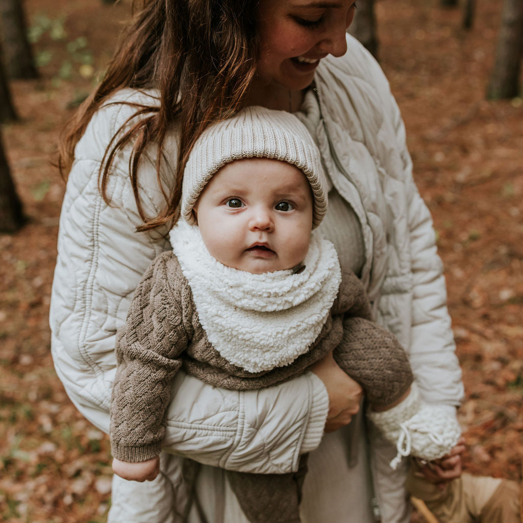 Bébé dans les bras de sa maman lors d'une marche automnale portant sa tuque beige en tricot de Petit Coulou et son cache-cou en sherpa