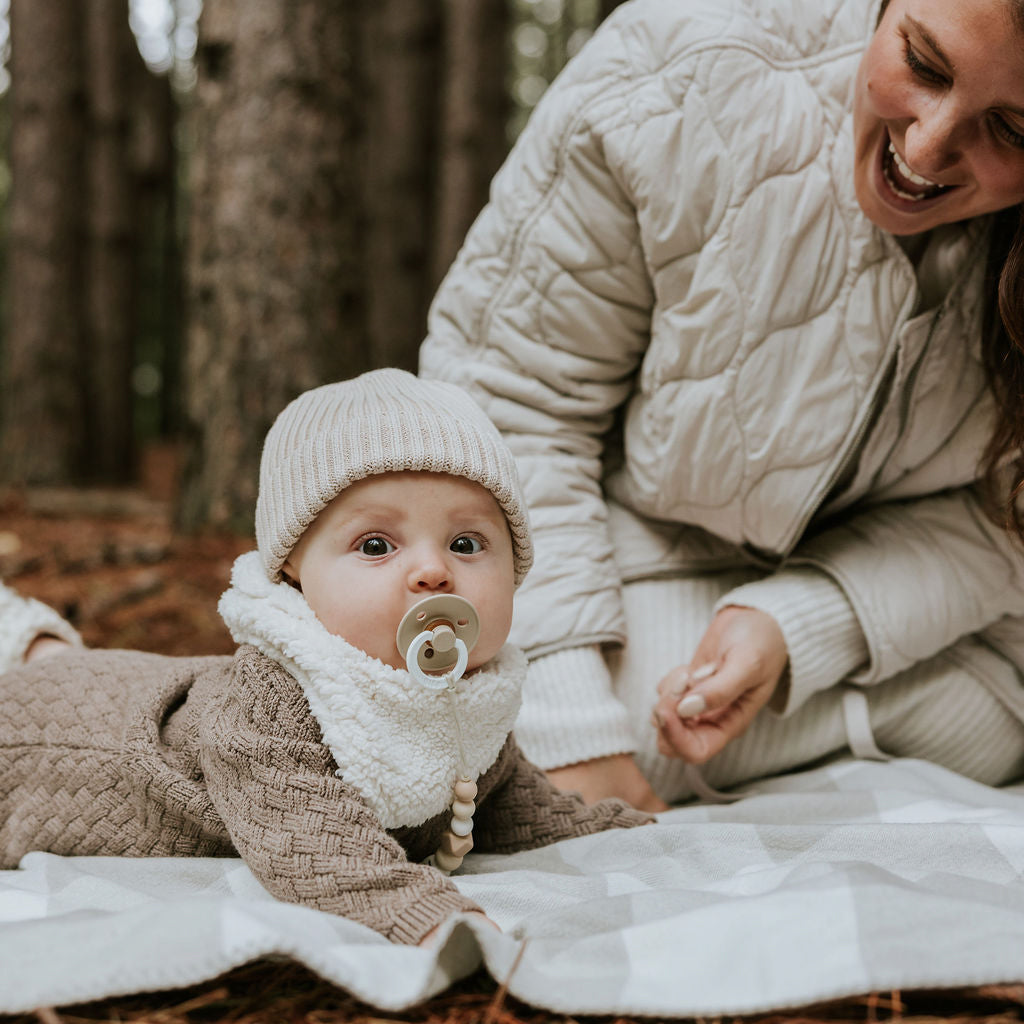 Bébé couché sur le ventre près de sa maman sur une couverte portant sa tque en tricot beige Petit Coulou et son cache-cou en sherpa