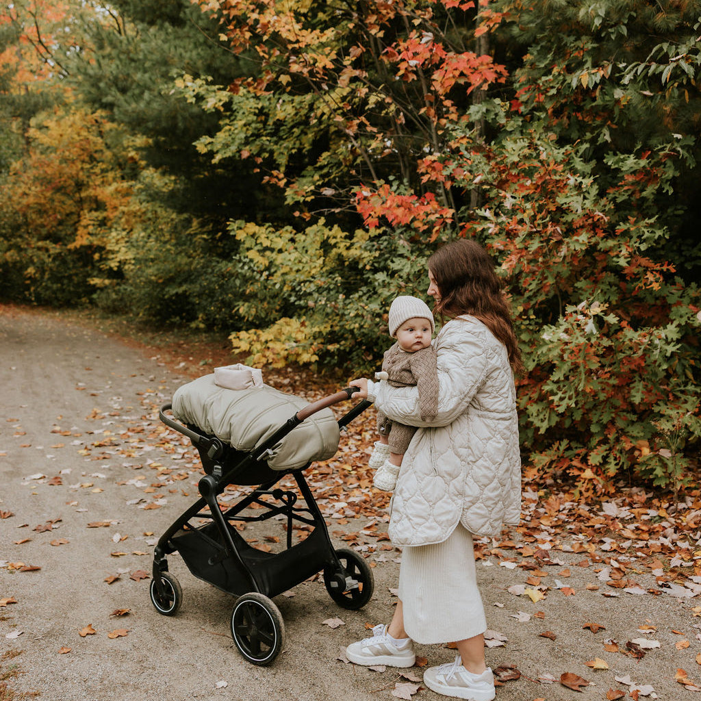 Bébé dans les bras de sa maman lors d'une marche automnale près de sa poussette avec coquille recouvert d'une housse légère lichen Petit Coulou