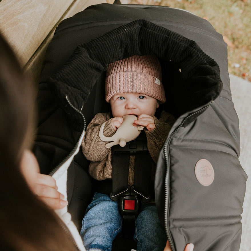 Bébé aux yeux bleus assis dans sa coquille recouvert d'une housse légère noire Petit Coulou et qui porte une petite tuque en tricot rose