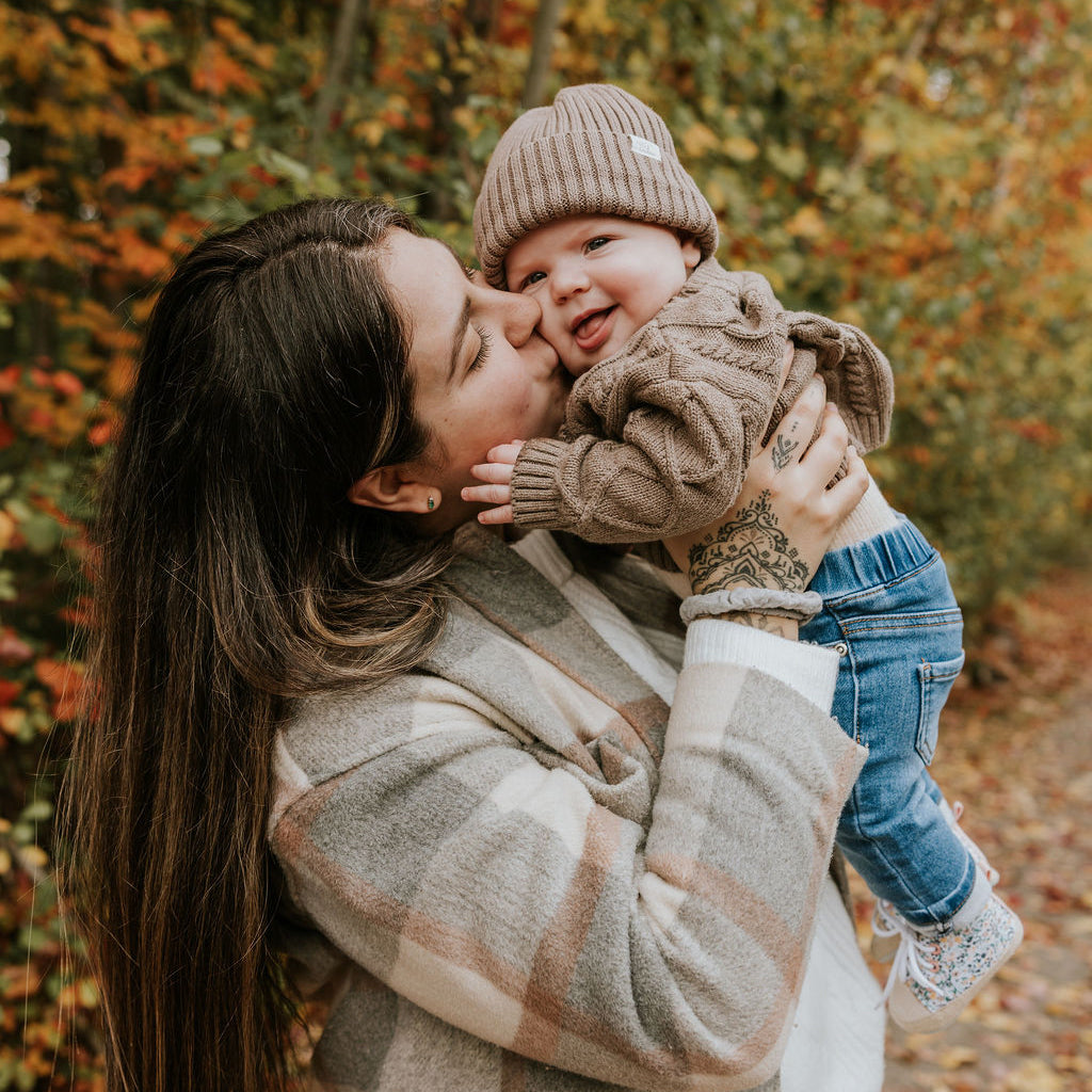 Mère qui donne un bisou sur la joue de son bébé qui porte une tuque en tricot taupe Petit Coulou