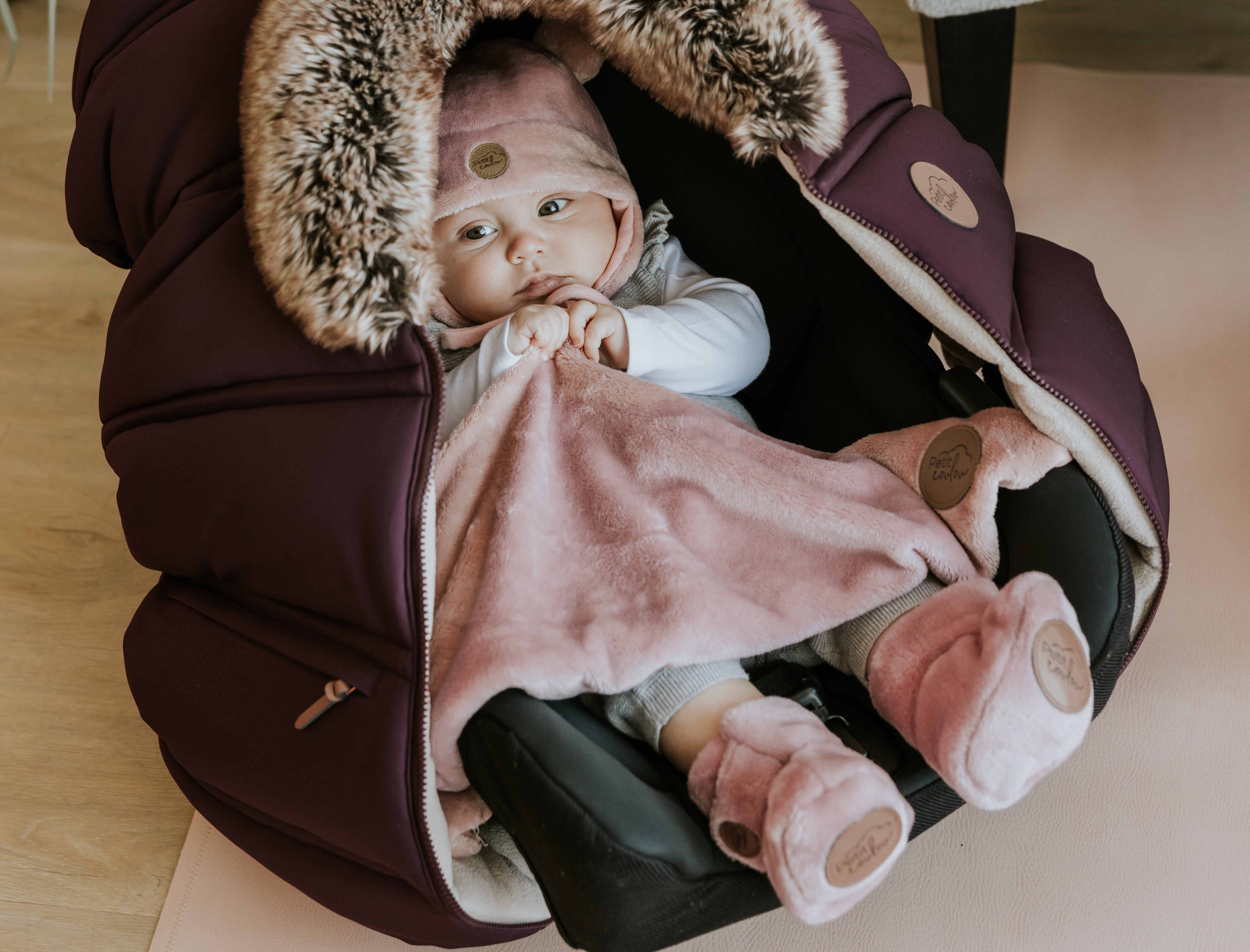 Bébé assis confortablement dans sa coquille recovert d'une housse d'hiver Petit Coulou couleur prune et qui porte ses accessoires en minky rose